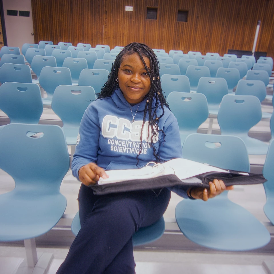 une élève qui sourit, avec son cartable à la main assise à l'auditorium de l'école